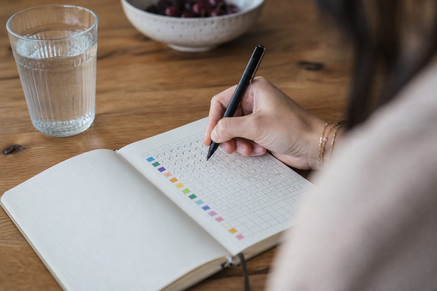 A hand holding a pen, ticking off a row of coloured habit boxes in an open paper tracker journal on a kitchen table, a glass of water nearby, natural daylight.