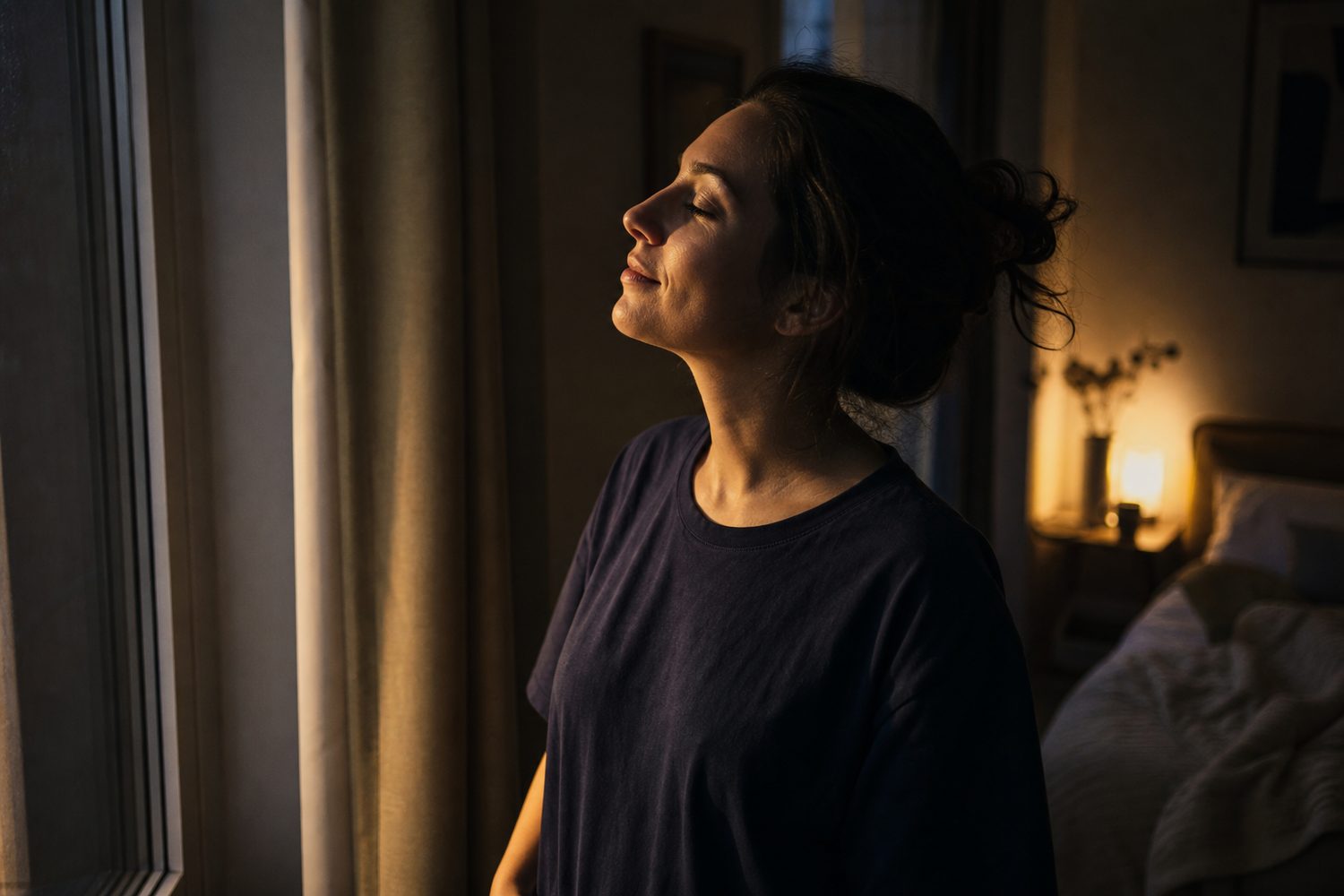 A woman standing by a window in the evening light with her eyes softly closed, taking a quiet breathing pause before bed
