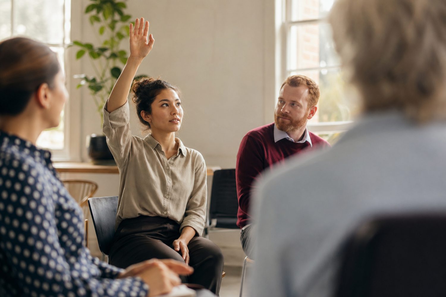 A young woman raising her hand to speak in a small bright meeting room, a slightly nervous but determined expression, warm daylight through a window, other people softly out of focus.