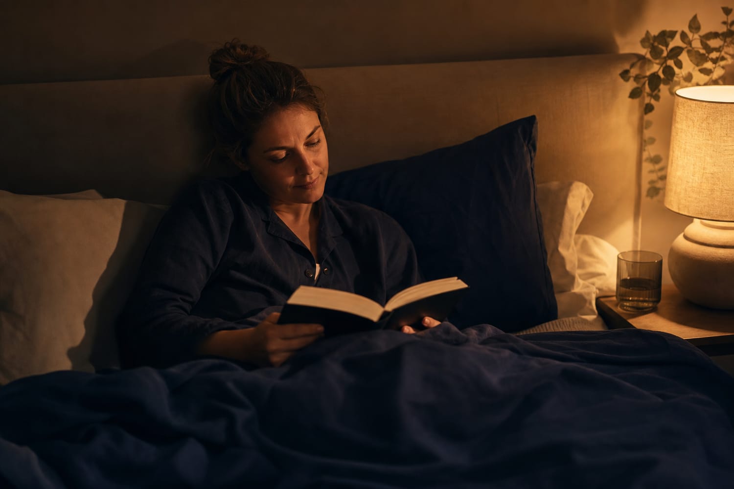 A woman reading a book in bed under warm, soft lamplight, relaxed and sleepy as she winds down for the night.